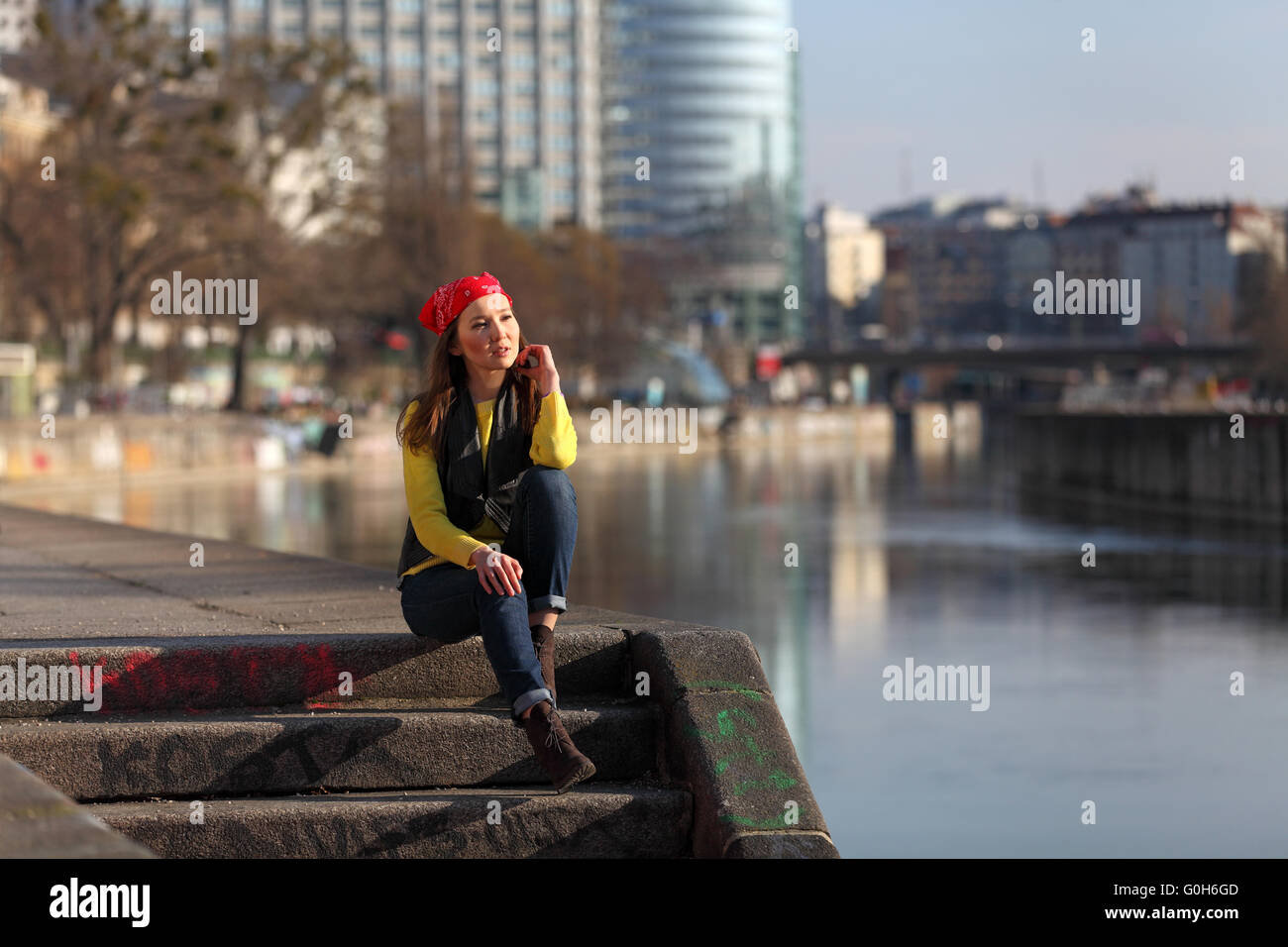 Beautiful Asian woman in yellow sweater and with red headscarf sitting at a river in a European city Stock Photo