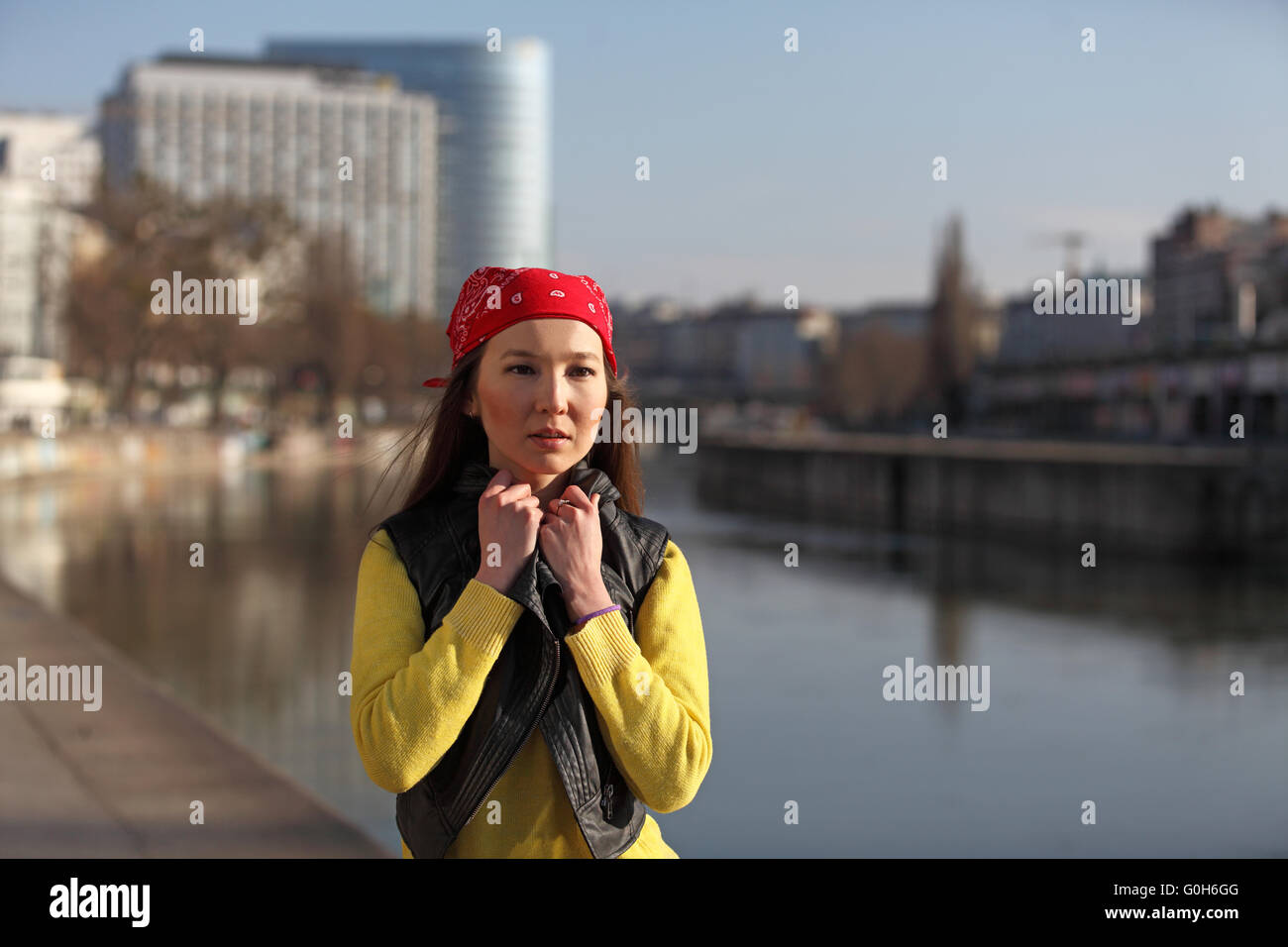 Beautiful Asian woman in yellow sweater and with red headscarf standing at a river in a European cit Stock Photo