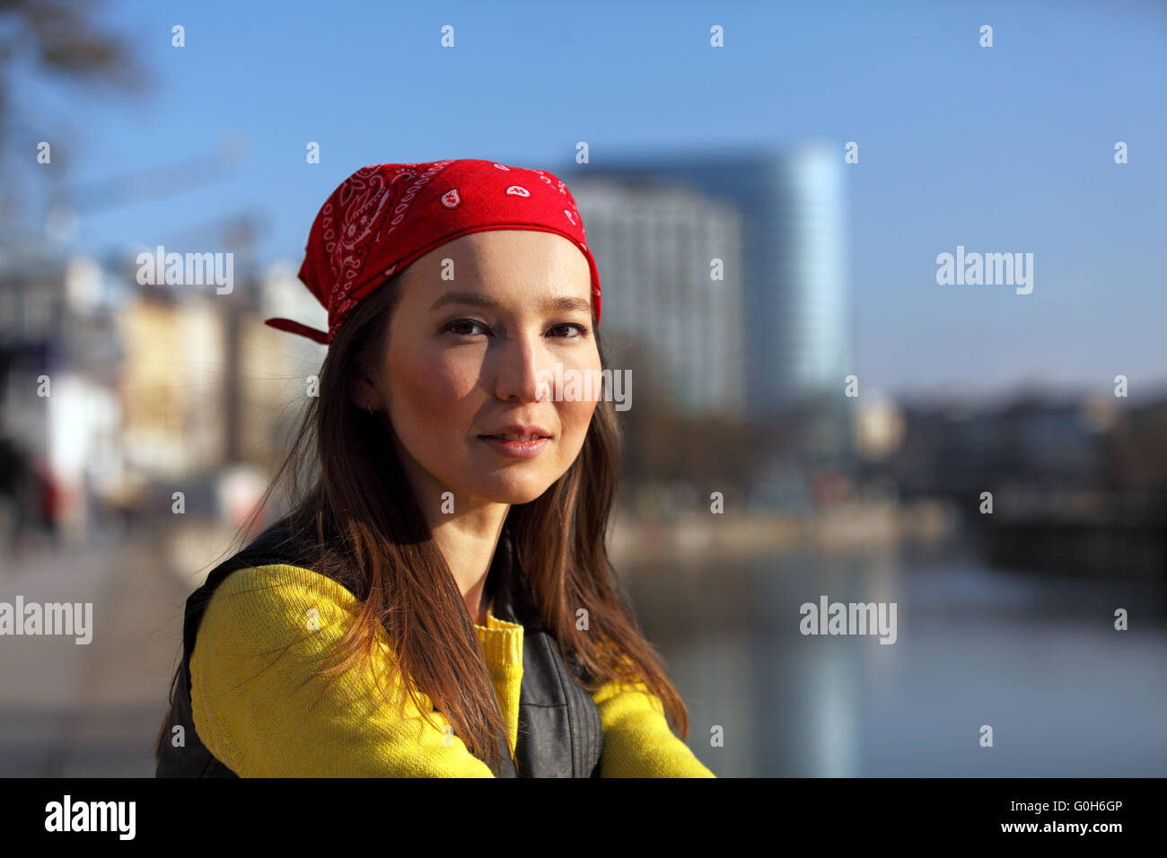 Beautiful Asian woman in yellow sweater and with red headscarf sitting at a river in a European city Stock Photo
