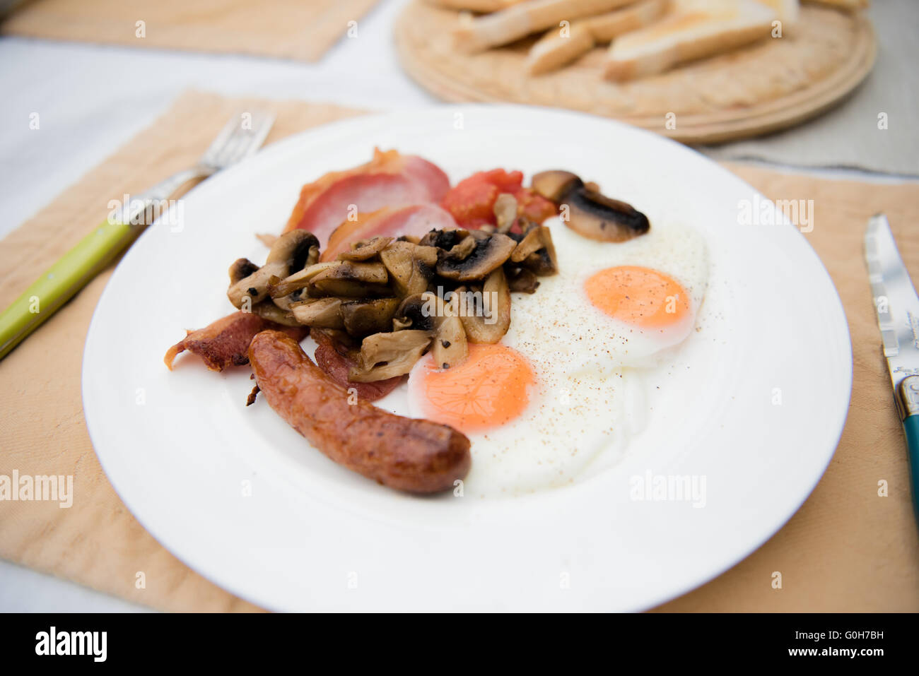 Full English breakfast on a white pate with two eggs, sausage, bacon, mushrooms, toast and brown