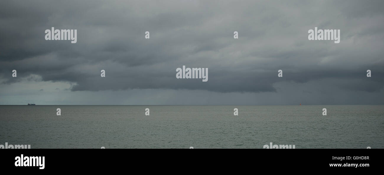 Container ship on horizon sailing toward storm cloud with sheets of rain falling onto flat calm green sea Stock Photo