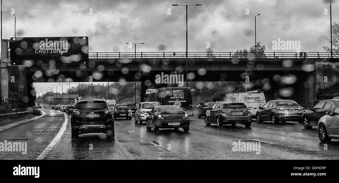 Queuing traffic on motorway with overhead matrix sign stating queue caution slightly blurred by raindrops on windscreen Stock Photo
