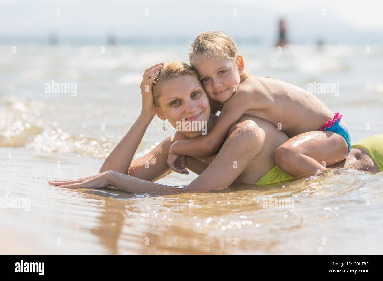 Mom and sits on her back the baby lying in the water on the sandy beach and happily look into the frame Stock Photo