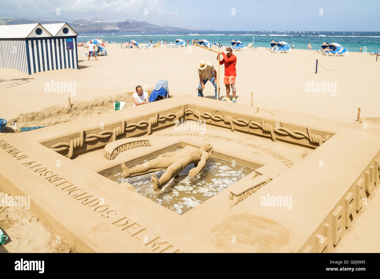 Refugee crisis sand sculpture, Las Palmas, Gran Canaria, Canary Islands, Spain, 1st May 2016. Sand sculptor/artist Etual Ojeda (centre) creates a stuning sand sculpture on the city beach, paying tribute to refugees who have lost their lives on treacherous sea crossings. The intricate work shows a person floating on water surrounded by barbed wire with the words 'Imagine there`s no countries' above his head. Below his feet the words 'Homenaje a los refugiados del mundo'. (tribute to the world`s refugees). Credit:  Alan Dawson News/Alamy Live News Stock Photo