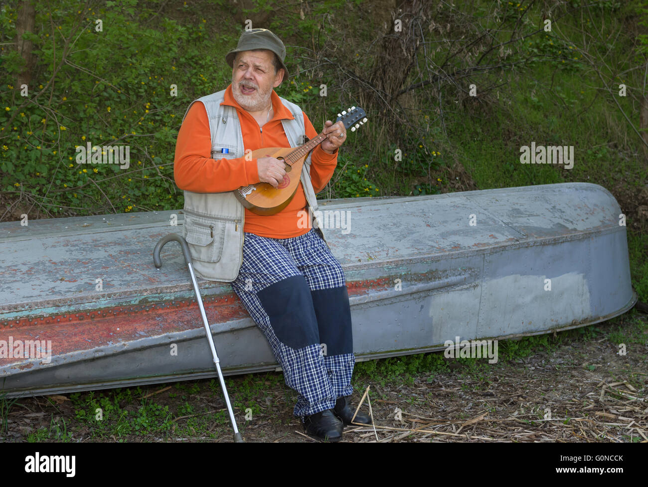 Senior man sitting on an old reversed boat and singing playing himself with mandolin Stock Photo