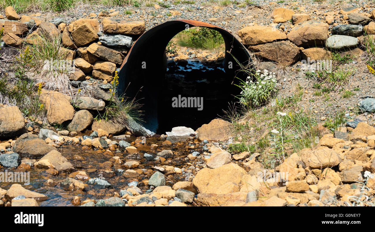 Looking through rusted corrugated culvert pipe embedded in rocky ground with water flowing from small stream. Stock Photo