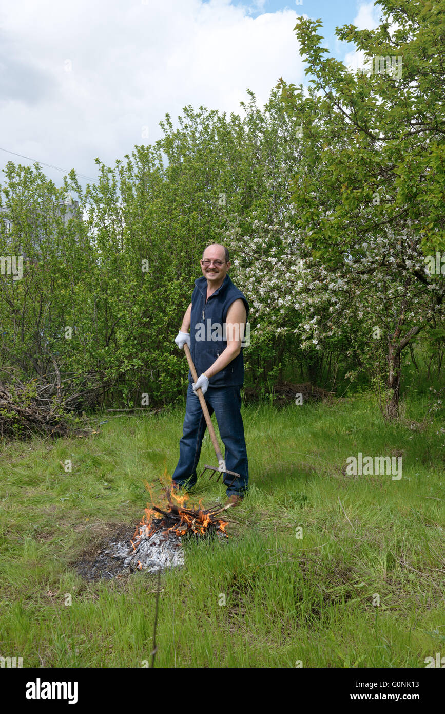 Bald mature man is standing with rake near small bonfire on bright ...