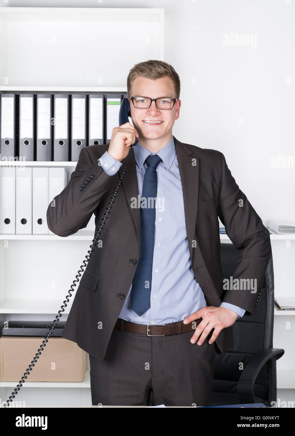 Young smiling businessman with glasses is phoning while standing at the desk in the office. A shelf is in the background. The ma Stock Photo