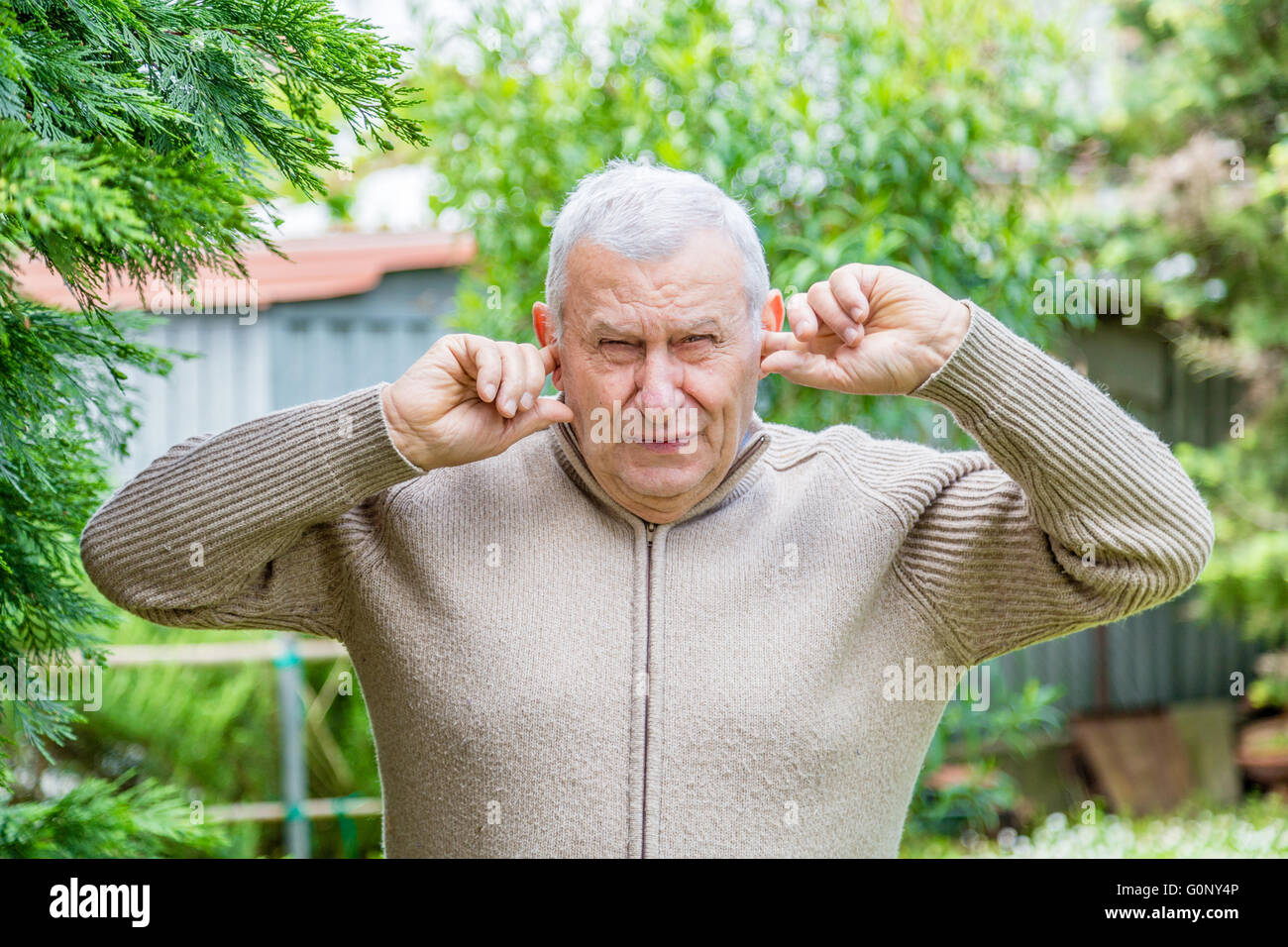 Elderly Caucasian in green garden covers his ears with his fingers not to hear Stock Photo