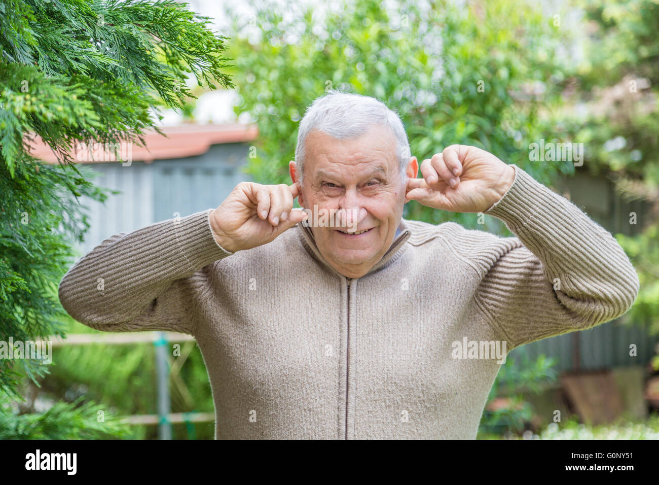 Elderly Caucasian in green garden covers his ears with his fingers not to hear Stock Photo
