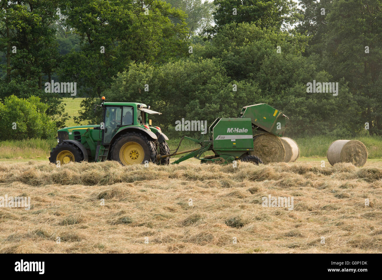 Great Ouseburn, North Yorkshire, GB, UK - round bale dropping & falling from a baler pulled by a green farm tractor working in a field, making silage. Stock Photo