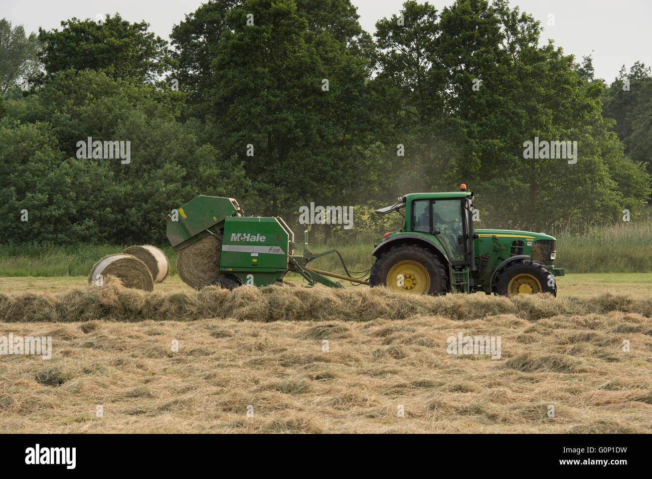 Great Ouseburn, North Yorkshire, GB - cylindrical bale is falling from round baler pulled by farm tractor driving & working in a field, making silage. Stock Photo