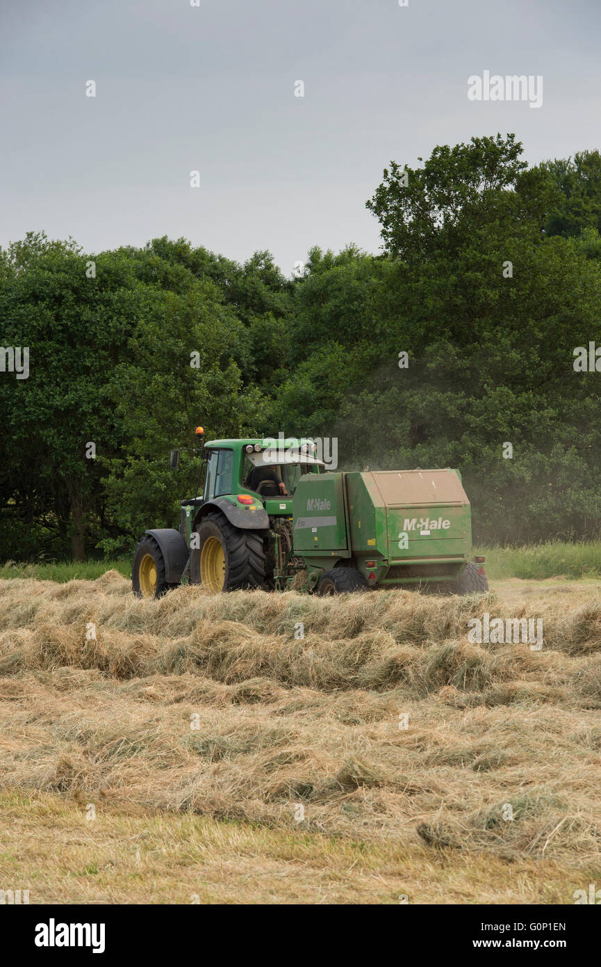 Hay or silage making - green, farm tractor pulling round baler, driving & working hard in field at Great Ouseburn, North Yorkshire, England, UK. Stock Photo
