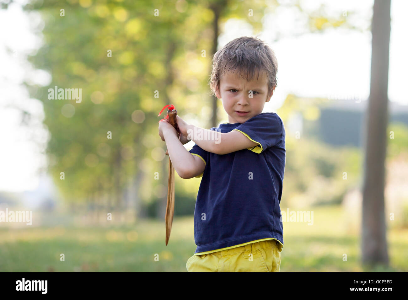 Angry little boy, holding sword, glaring with a mad face at the camera, outdoors in the park Stock Photo