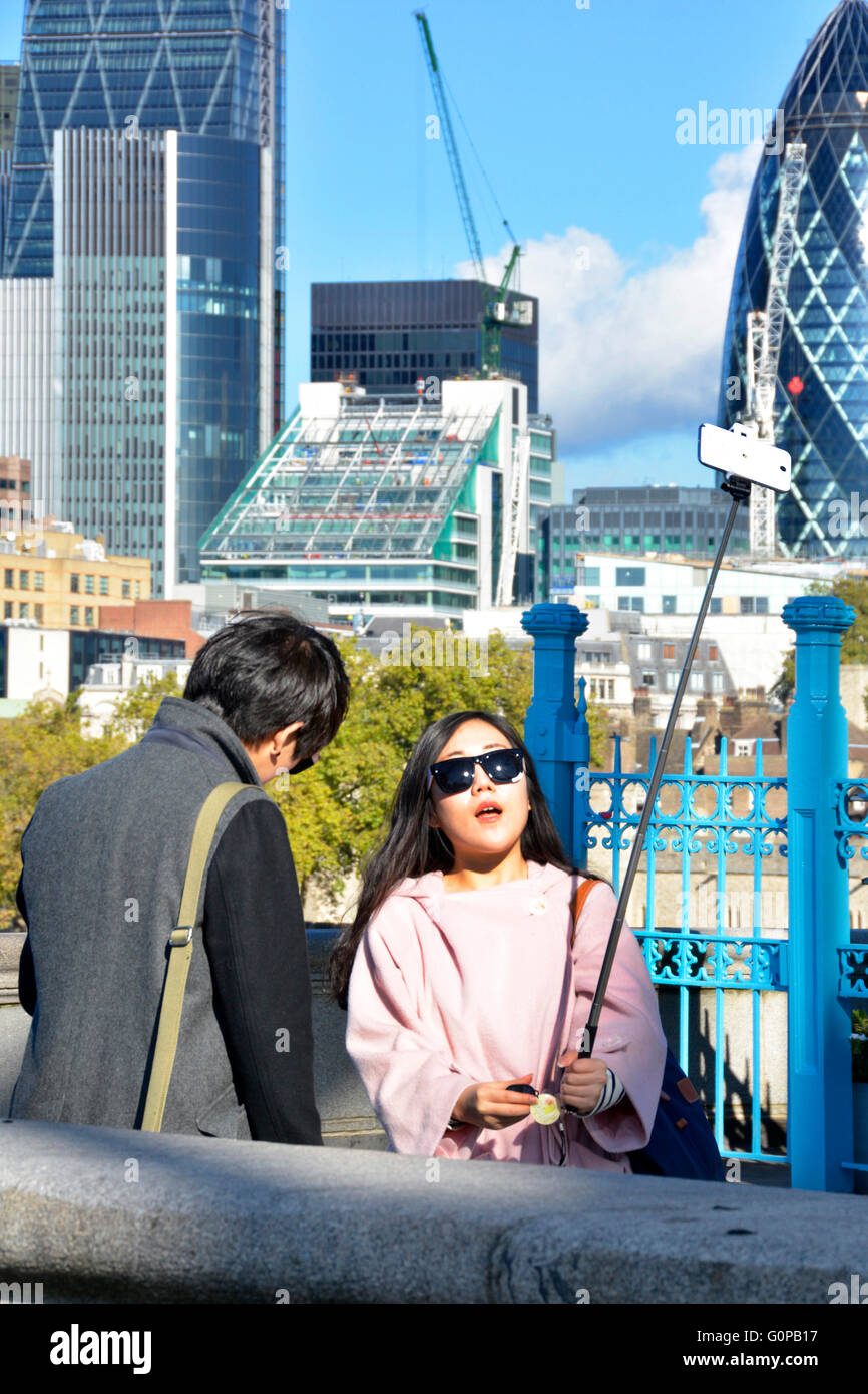 Woman tourist on Tower Bridge pavement composing a photo using selfie stick and smart phone with City of London skyline view beyond England UK Stock Photo