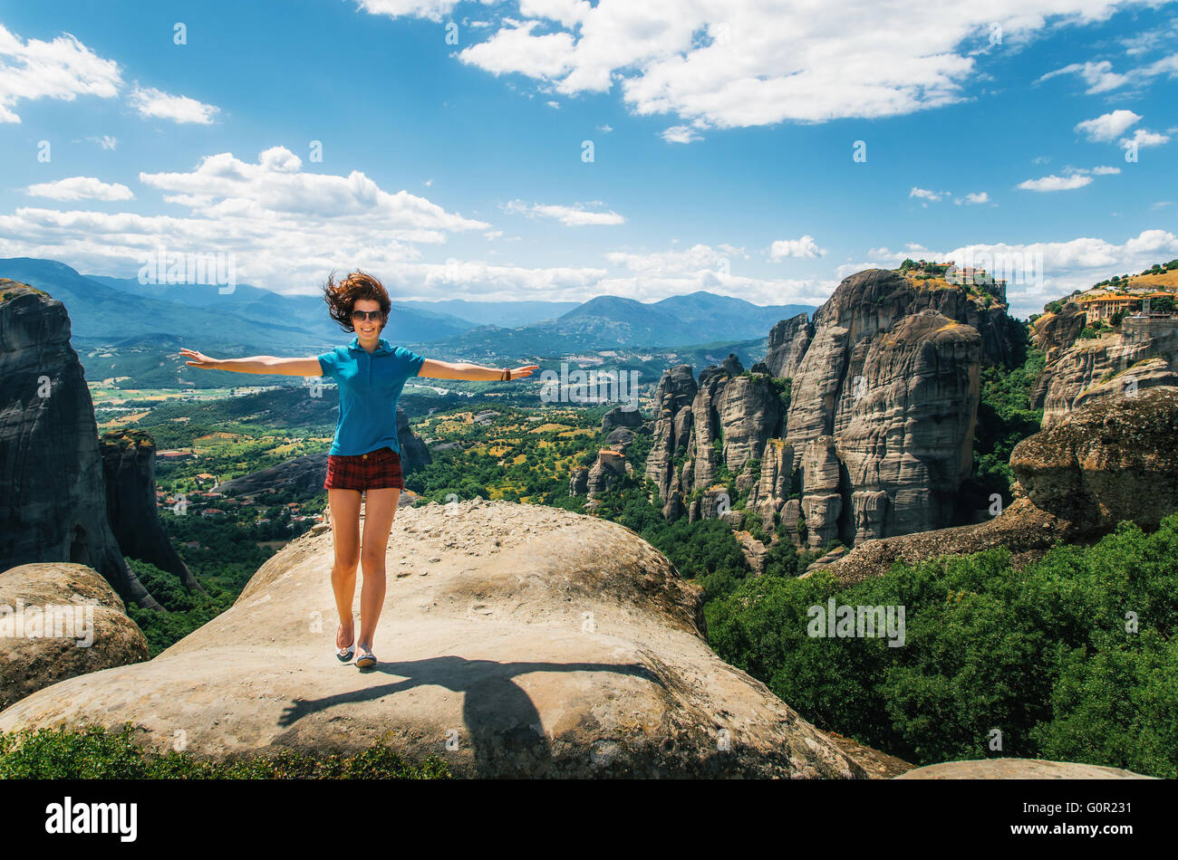 Young happy woman stands back to the rock with raised hands. Traveler enjoying the landscape, Meteora, Greece Stock Photo