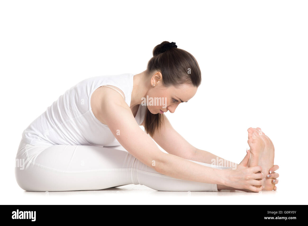 Sporty beautiful young woman in white sportswear sitting in seated forward bend pose, doing paschimothanasana posture Stock Photo