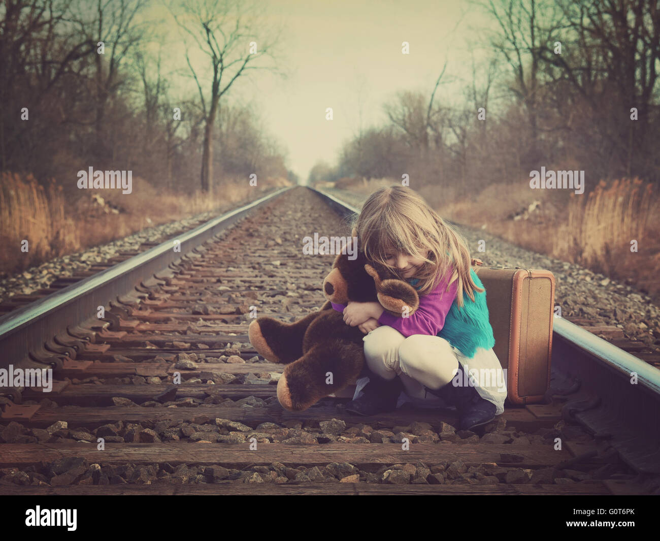 A little girl is sitting on an old train track outside and hugging a teddy bear for a vintage memory or travel concept. Stock Photo