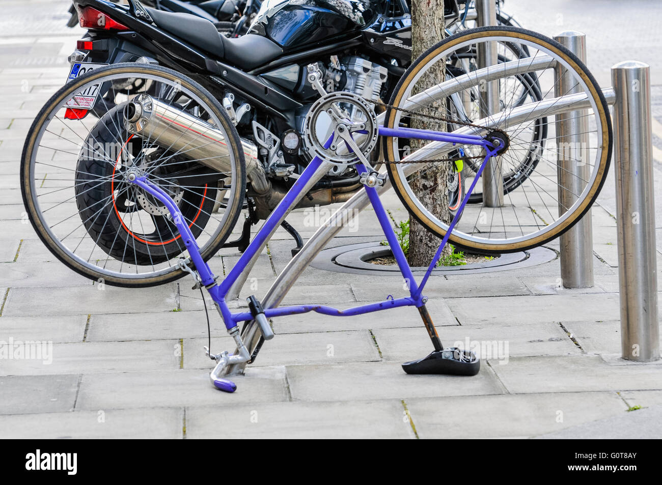 An old, wrecked bicycle is turned upside-down and locked to a bicycle rack on a pavement. Stock Photo