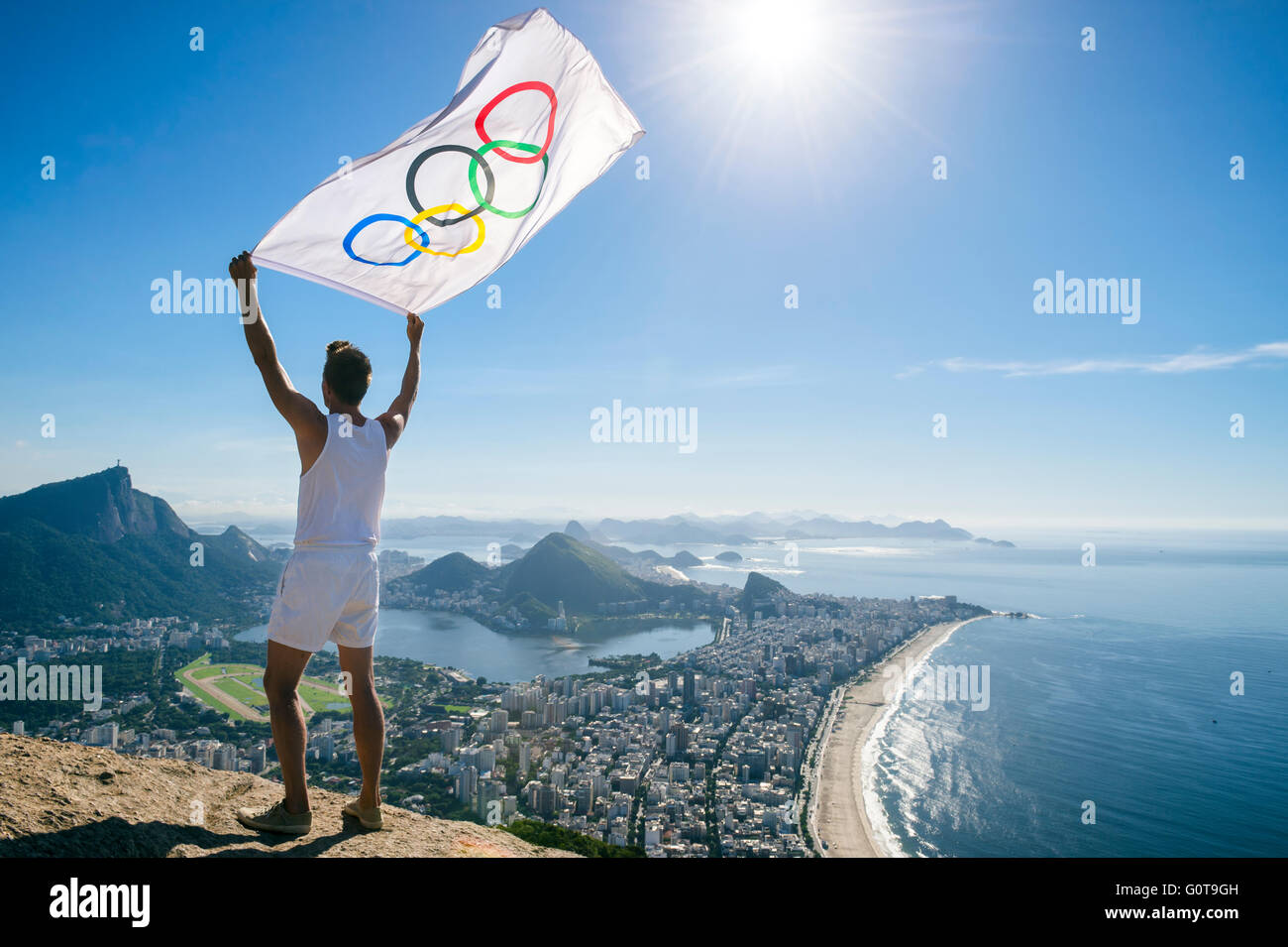 RIO DE JANEIRO - MARCH 21, 2016: Athlete stands holding Olympic flag above a city skyline view of the beaches and Corcovado. Stock Photo