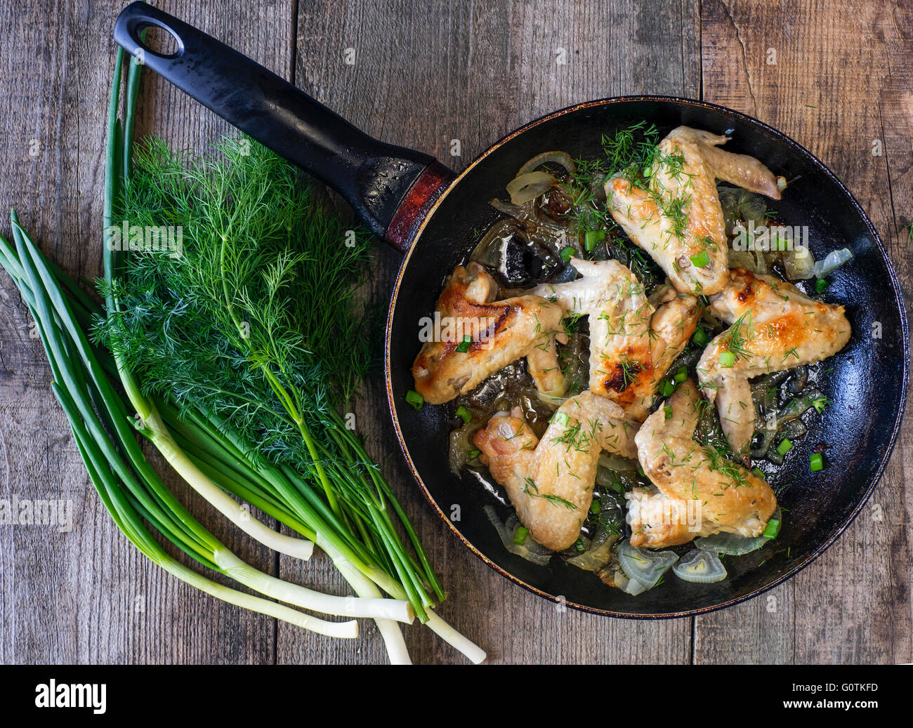 Fried chicken wings in frying pan with herbs and spring onions on wooden table Stock Photo
