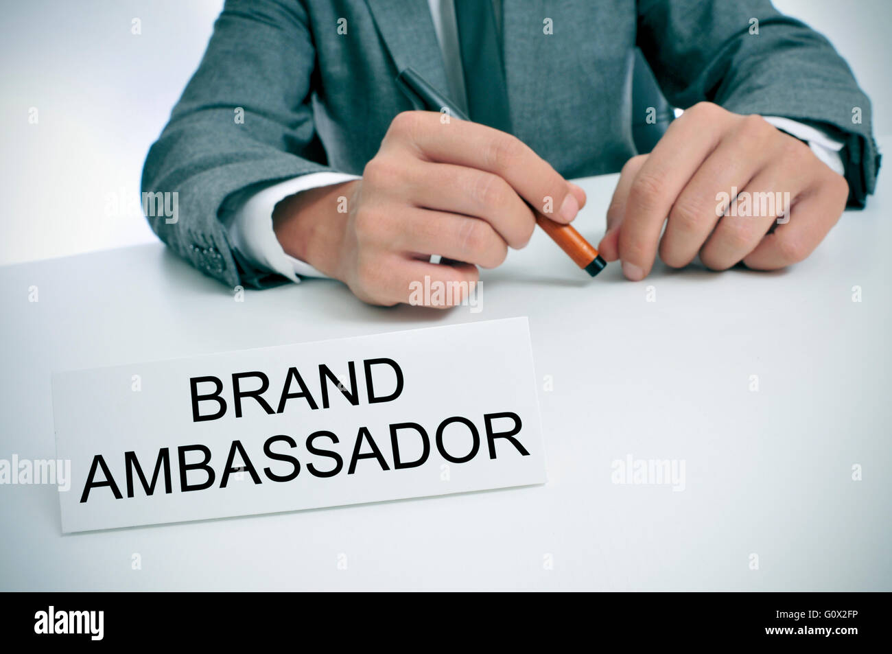 closeup of a young caucasian man wearing a gray suit sitting at his office desk, with a name plate in front of him with the text Stock Photo