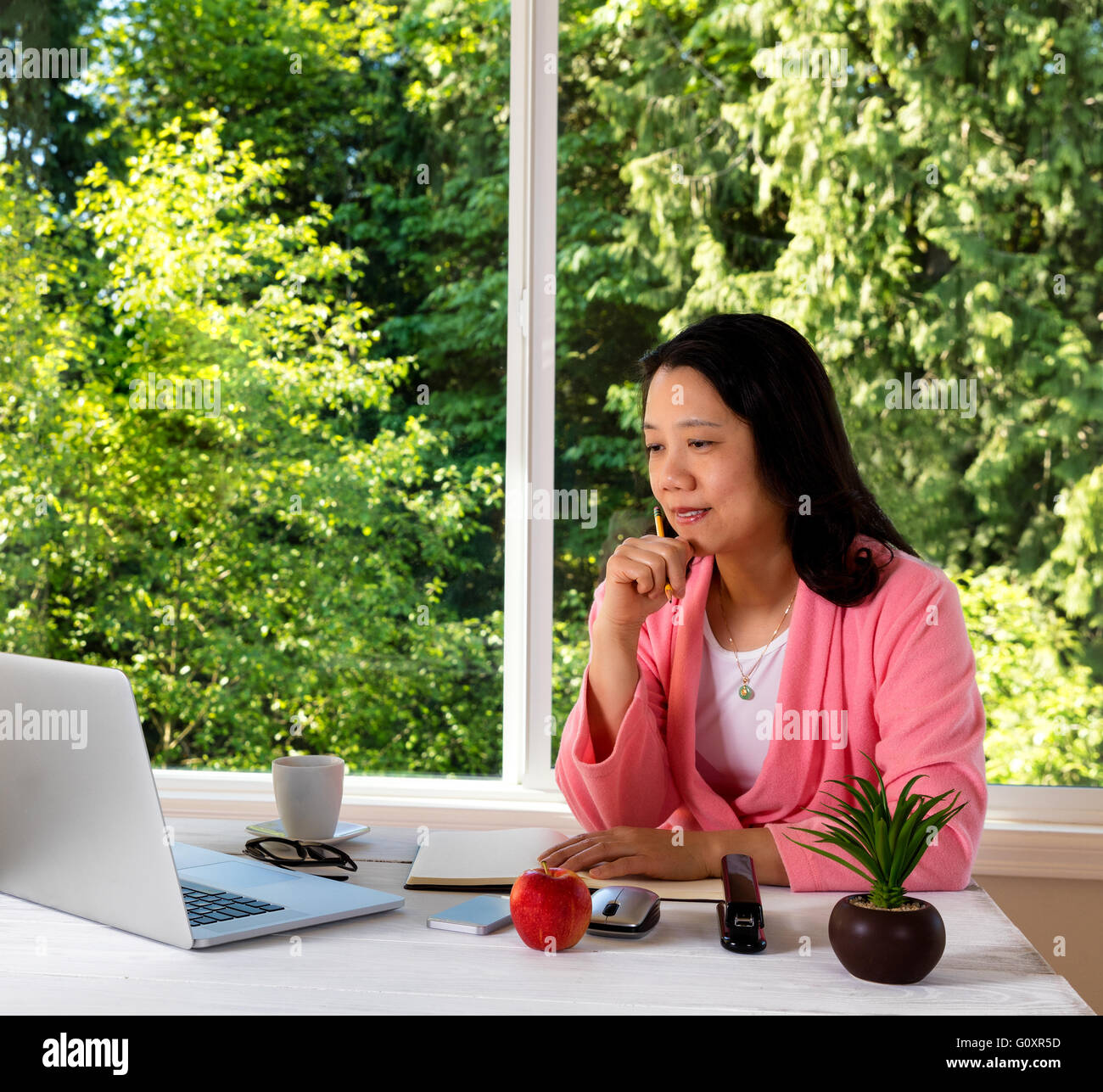 Mature woman, wearing pink bathrobe, working from home in front of large window with bright daylight and trees in background. Stock Photo