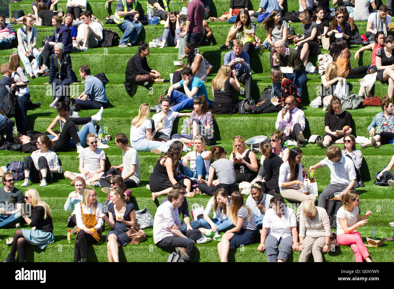 A large group of friends, co-workers and colleagues enjoying their lunch break in the sunshine on a grassy bank in the city. Stock Photo