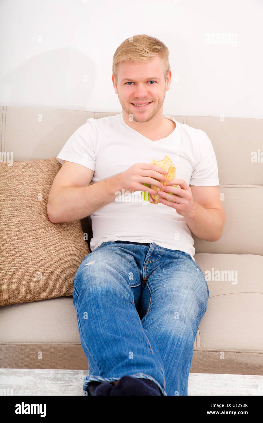 A young adult man eating a sandwich at home on the couch. Stock Photo