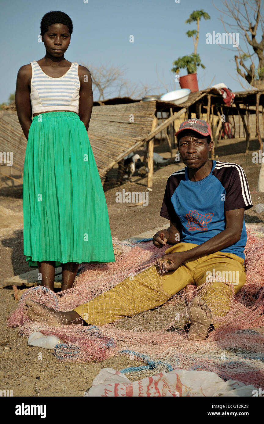 Fisherman sitting on the beach and mending a fishing net with his wife standing beside him in the village of Chembe, Malawi Stock Photo