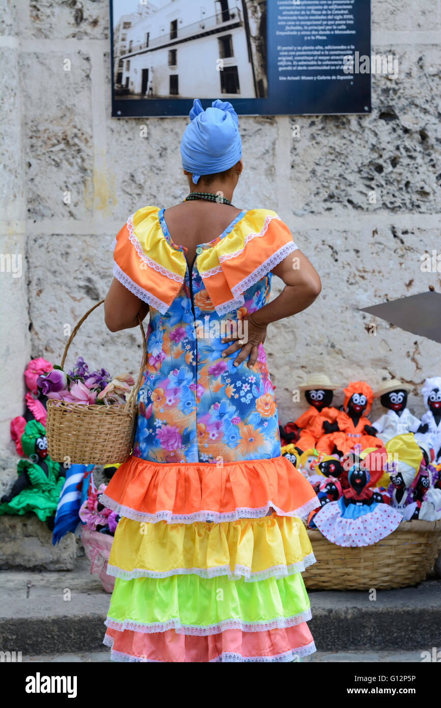 A flower seller wearing traditional costume looks at cloth dolls on sale in the Plaza de la Catedral, Old Havana, Havana, Cuba Stock Photo