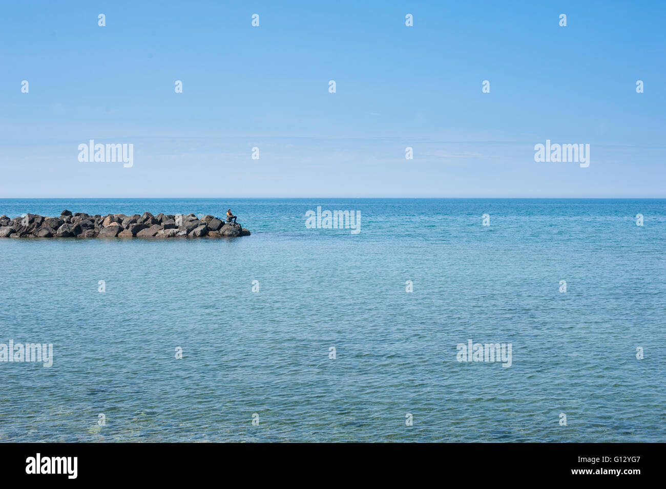 Fisherman alone on rock and large blue sea landscape in sunny summer day Stock Photo