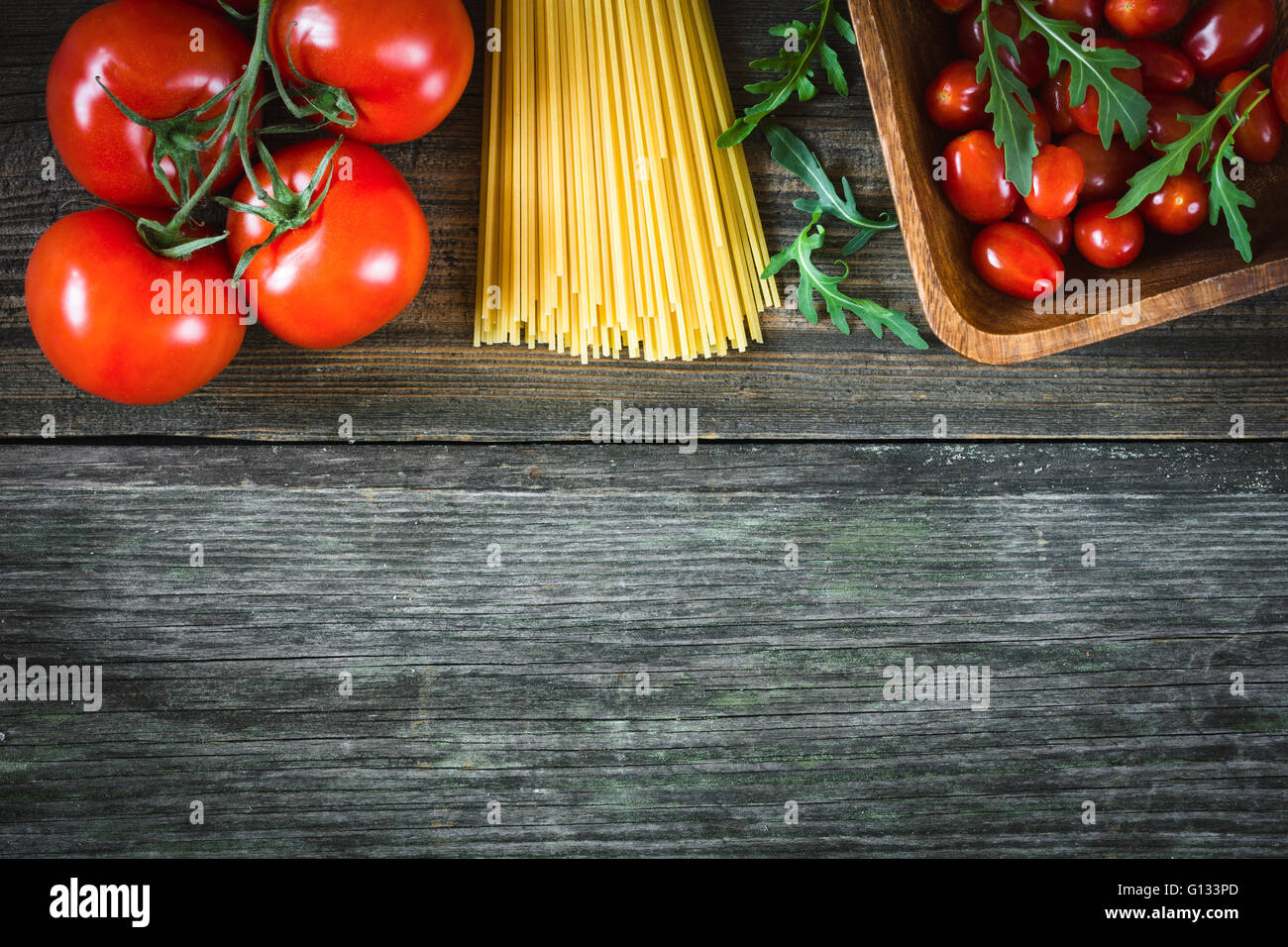 Italian food background / cooking ingredients background. Dry pasta, tomatoes, olive oil and salad leaves on wooden backdrop Stock Photo