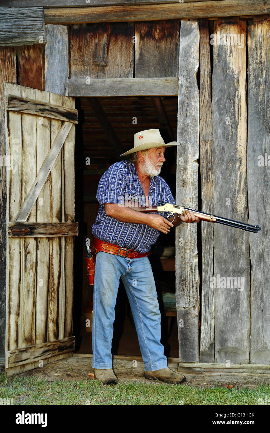 A mature aged cowboy leans against a wooden barn with rifle and pistol at the ready. Stock Photo