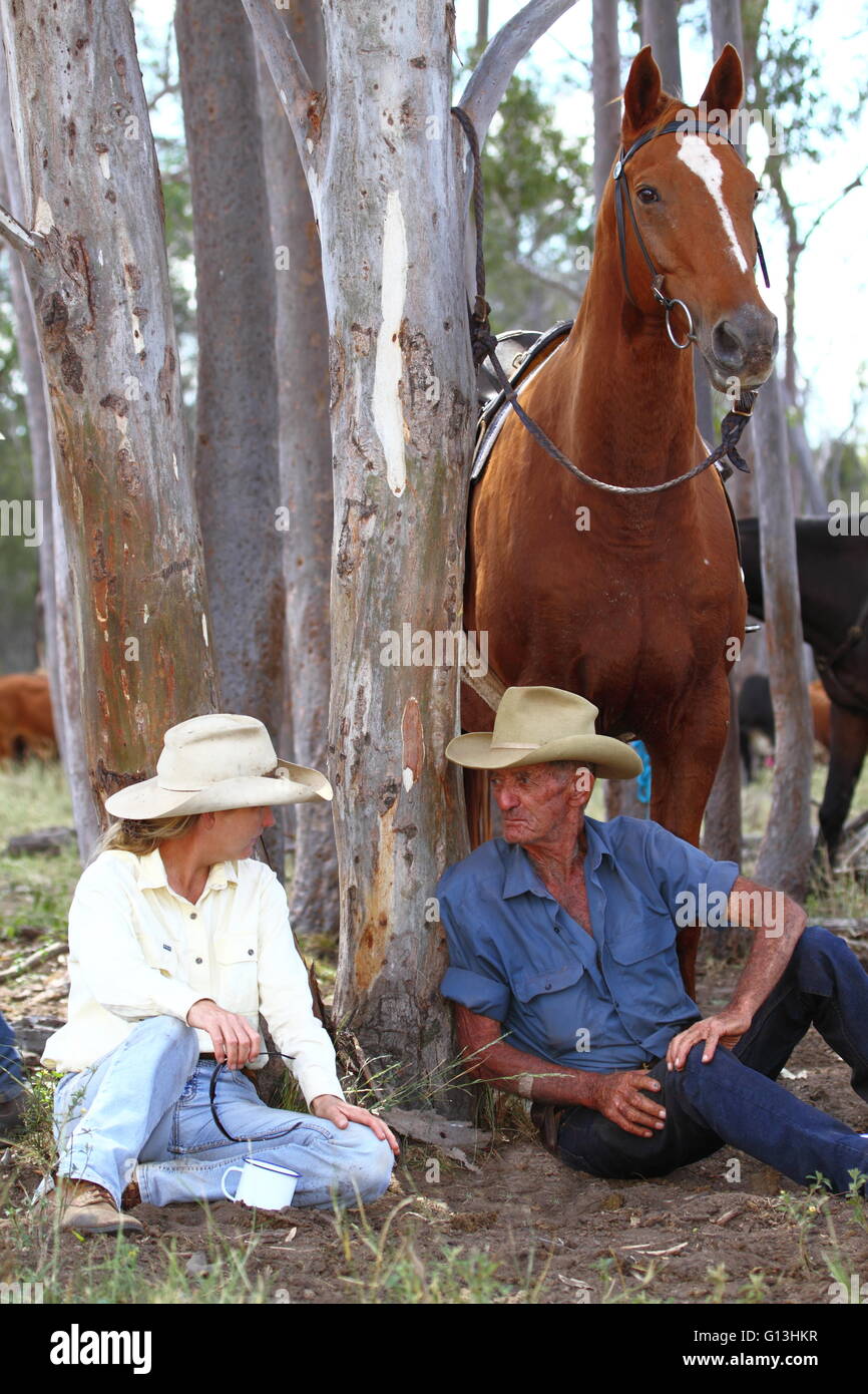 A cowgirl in her 30's chats with an older cattleman cowboy in his 80's as they rest against a tree with horses in background Stock Photo