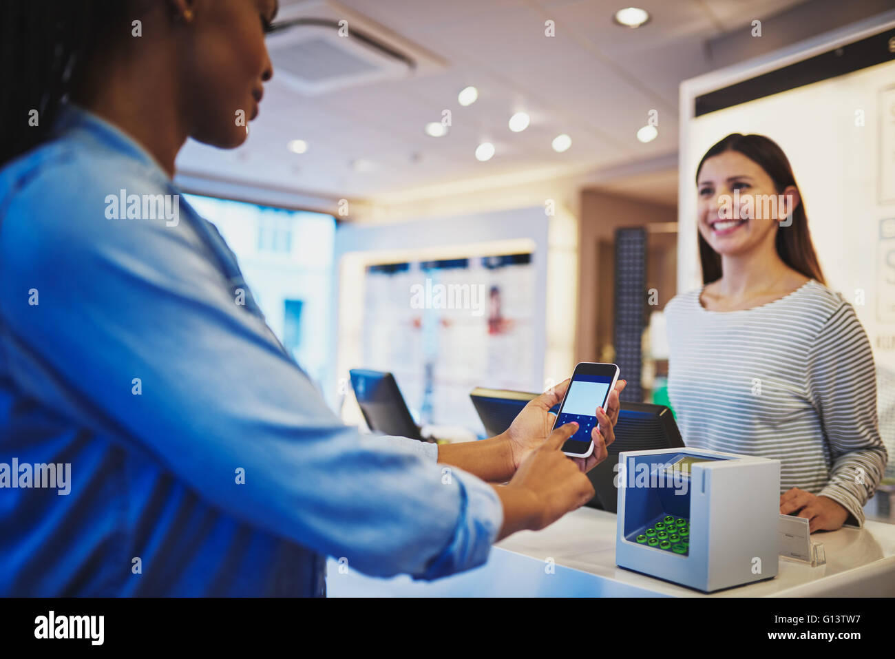 Smiling cashier in striped blouse watching customer pay at register with phone in eyeglasses store Stock Photo