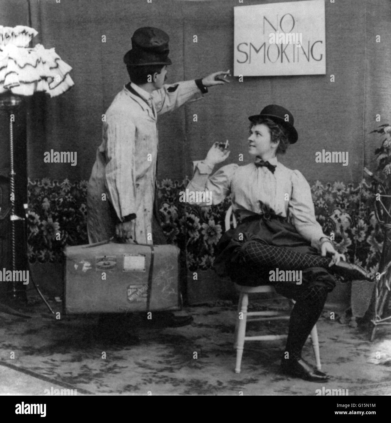 Humorous scene of man pointing to No Smoking sign above woman in derby and bloomers who is smoking cigarette. Bloomers is a word which has been applied to several types of divided women's garments for the lower body at various times. The costume was calle Stock Photo