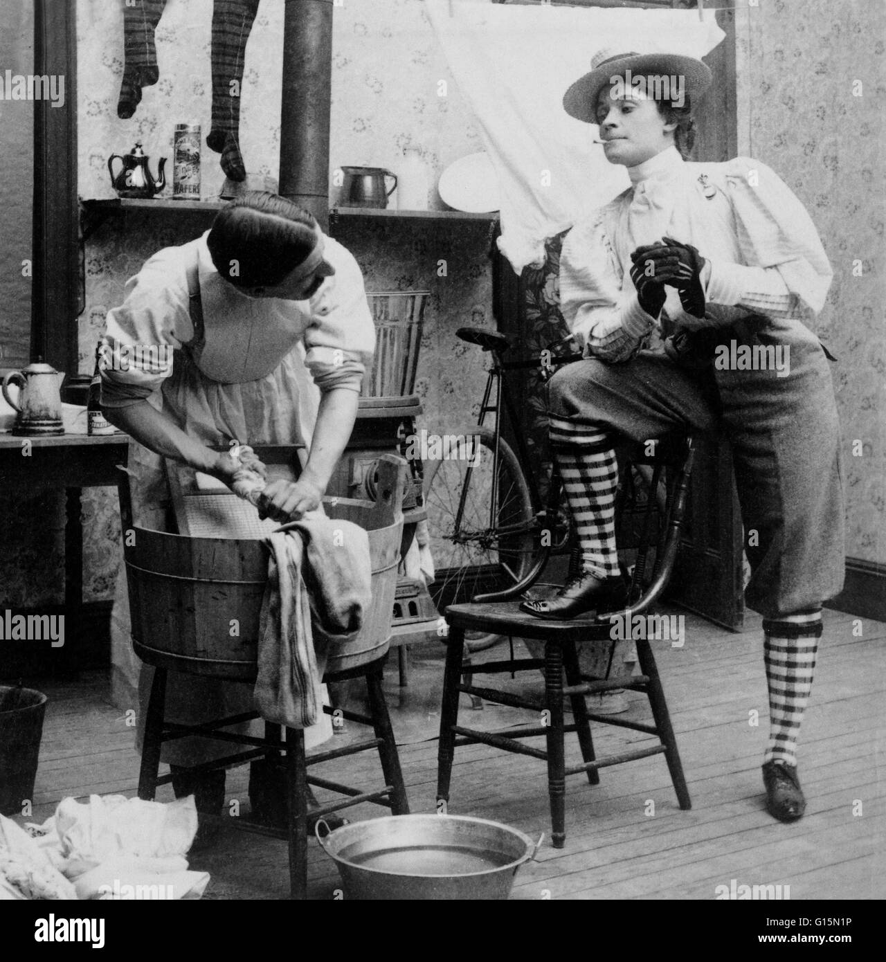 Woman in bloomers smoking cigarette and looking at man doing laundry, 1901. Bloomers is a word which has been applied to several types of divided women's garments for the lower body at various times. The costume was called the American Dress or Reform Cos Stock Photo