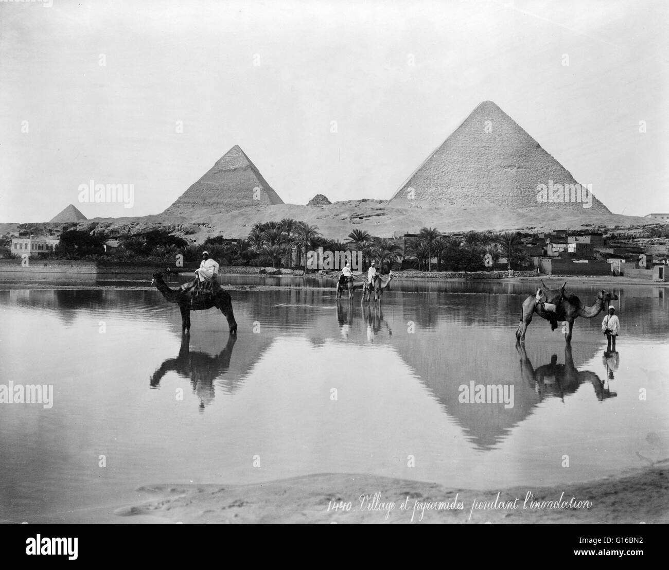Entitled: 'Village and pyramids during the flood-time, circa 189?'. shows men on camels and one man standing next to a camel in shallow flood water, with pyramids in the background. The Giza Necropolis (pyramids of Giza) is an archeological site on the Gi Stock Photo