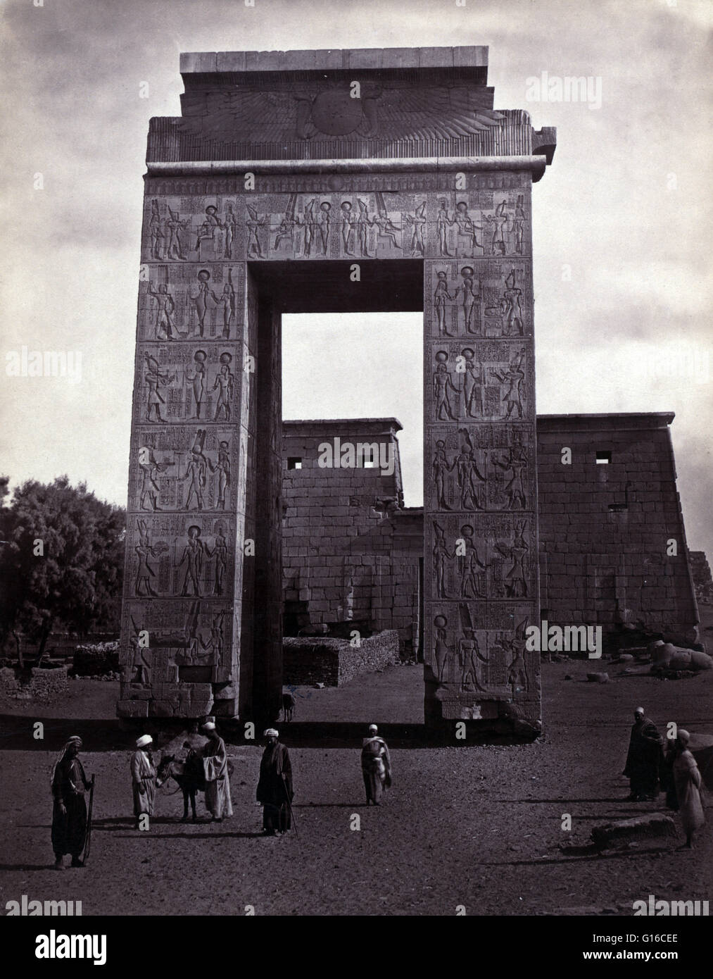 Entitled: "People near pylons, at Carnac." A pylon is a gateway to the inner part of an Ancient Egyptian temple. The Karnak Temple Complex comprises a vast mix of decayed temples, chapels, pylons, and other buildings. Building at the complex began during Stock Photo