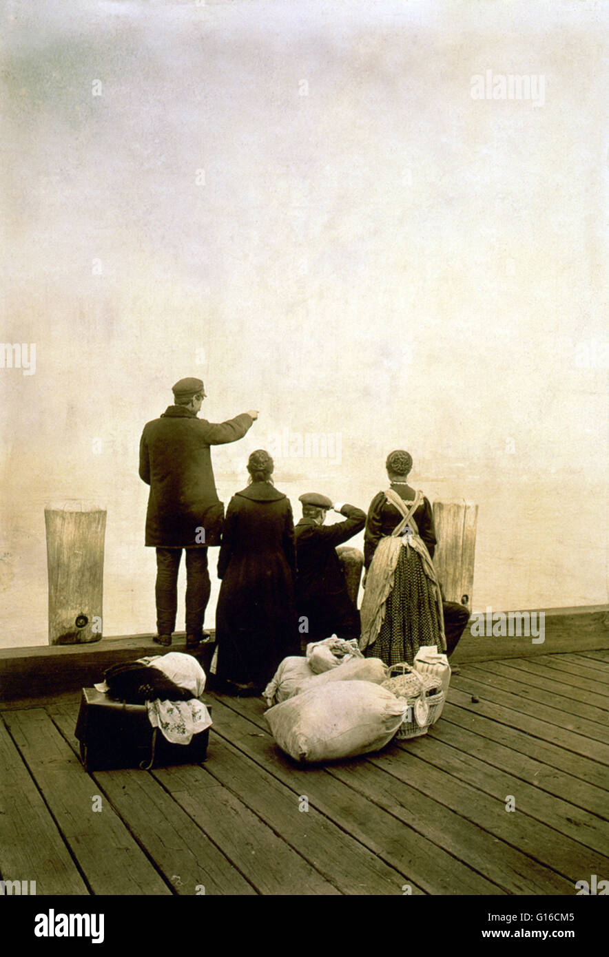 Entitled: 'Four immigrants and their belongings, on a dock, looking out over the water; view from behind.' In the 35 years before Ellis Island opened, over eight million immigrants arriving in New York had been processed by New York State officials at Cas Stock Photo