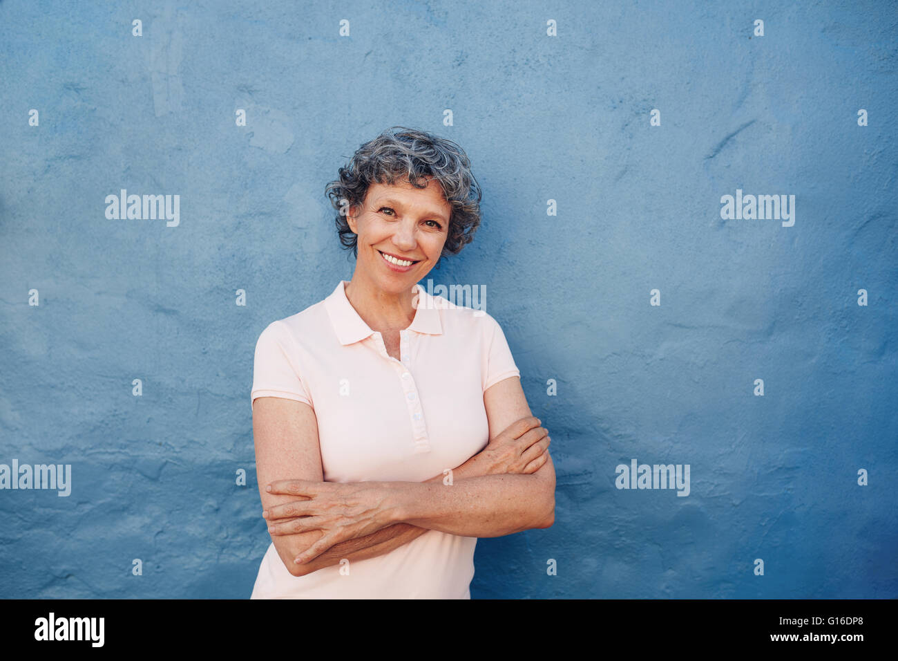 Portrait of attractive mature woman with her arms crossed standing against blue background. She is leaning to a blue wall with c Stock Photo