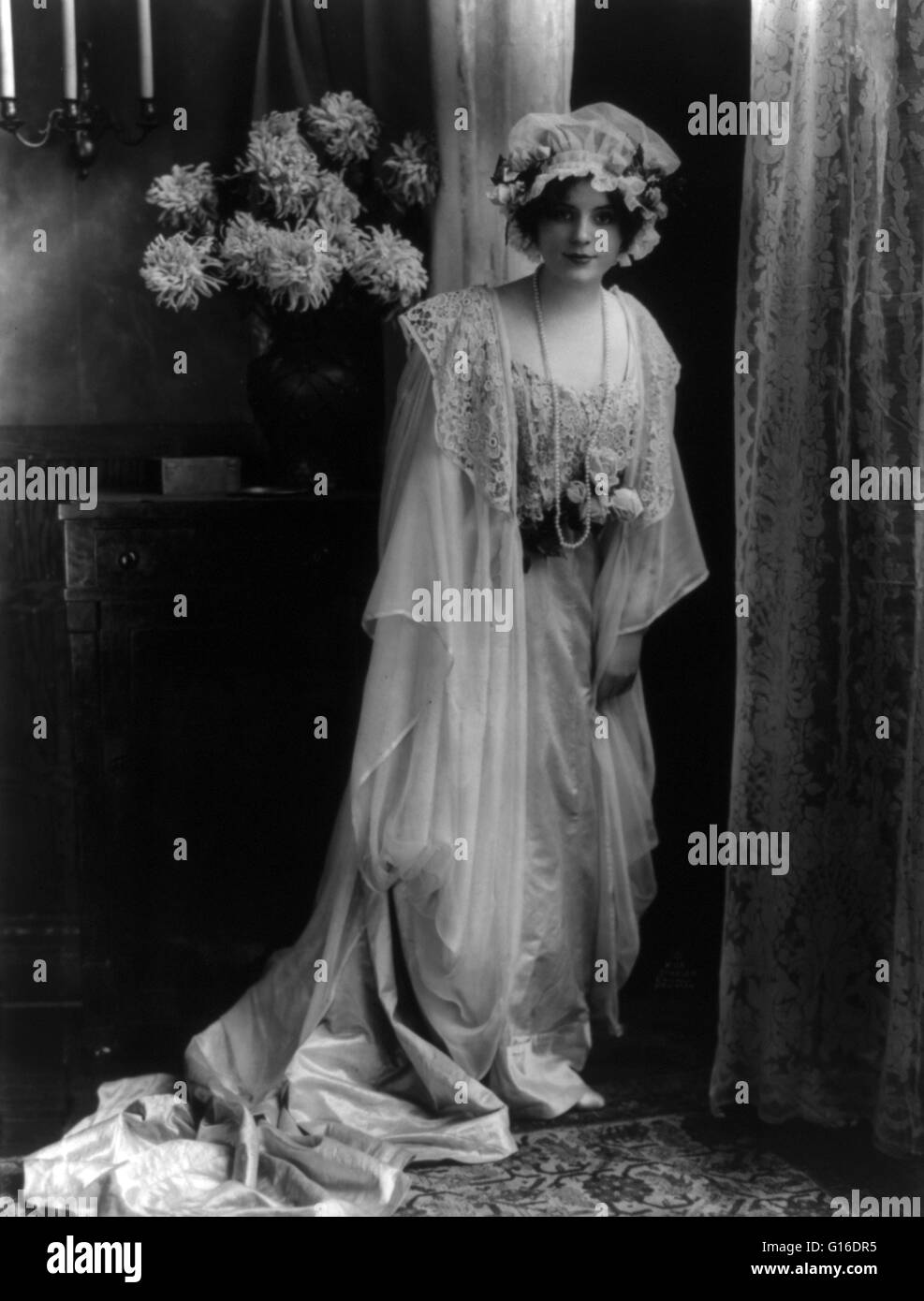 Young woman posed in lace gown and cap. A peignoir is a long outer garment for women which is frequently sheer and made of chiffon or another translucent fabric. Nightwear is clothing designed to be worn while sleeping. The style of nightwear worn may var Stock Photo