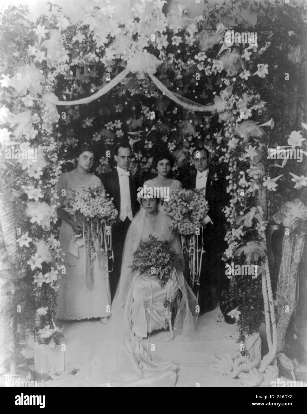 Entitled: 'Wedding party inside arbor of flower' shows an unidentified wedding party posed inside an arbor of flowers; bride seated in center. Wedding traditions and customs vary greatly between cultures, ethnic groups, religions, countries, and social cl Stock Photo