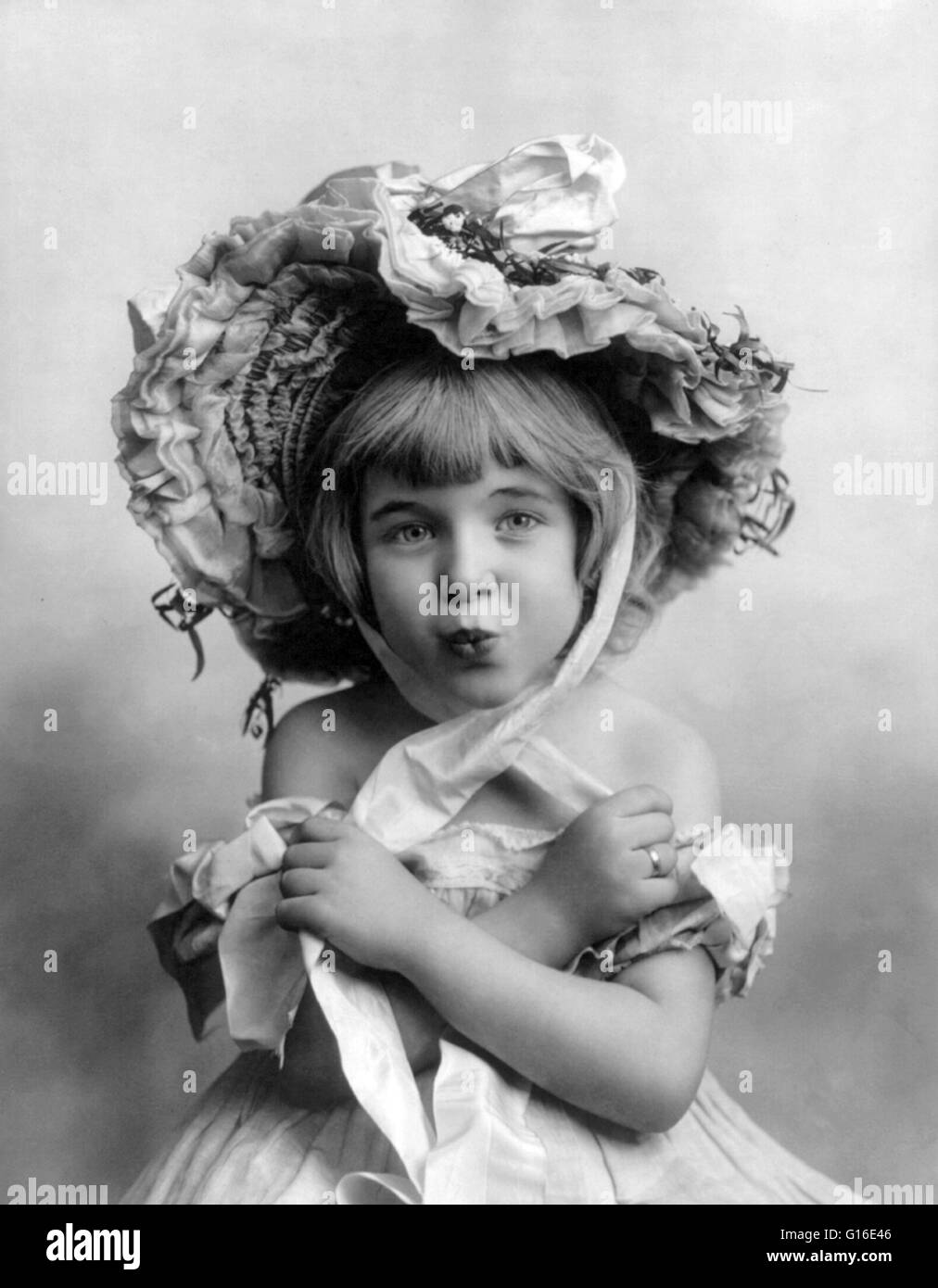 Entitled: 'Little girl, wearing bonnet, with arms folded and lips puckered.' An Easter bonnet is a type of hat that women and girls wear to Easter services, and in the Easter parade following it. Ladies purchased new and elaborate designs for particular c Stock Photo