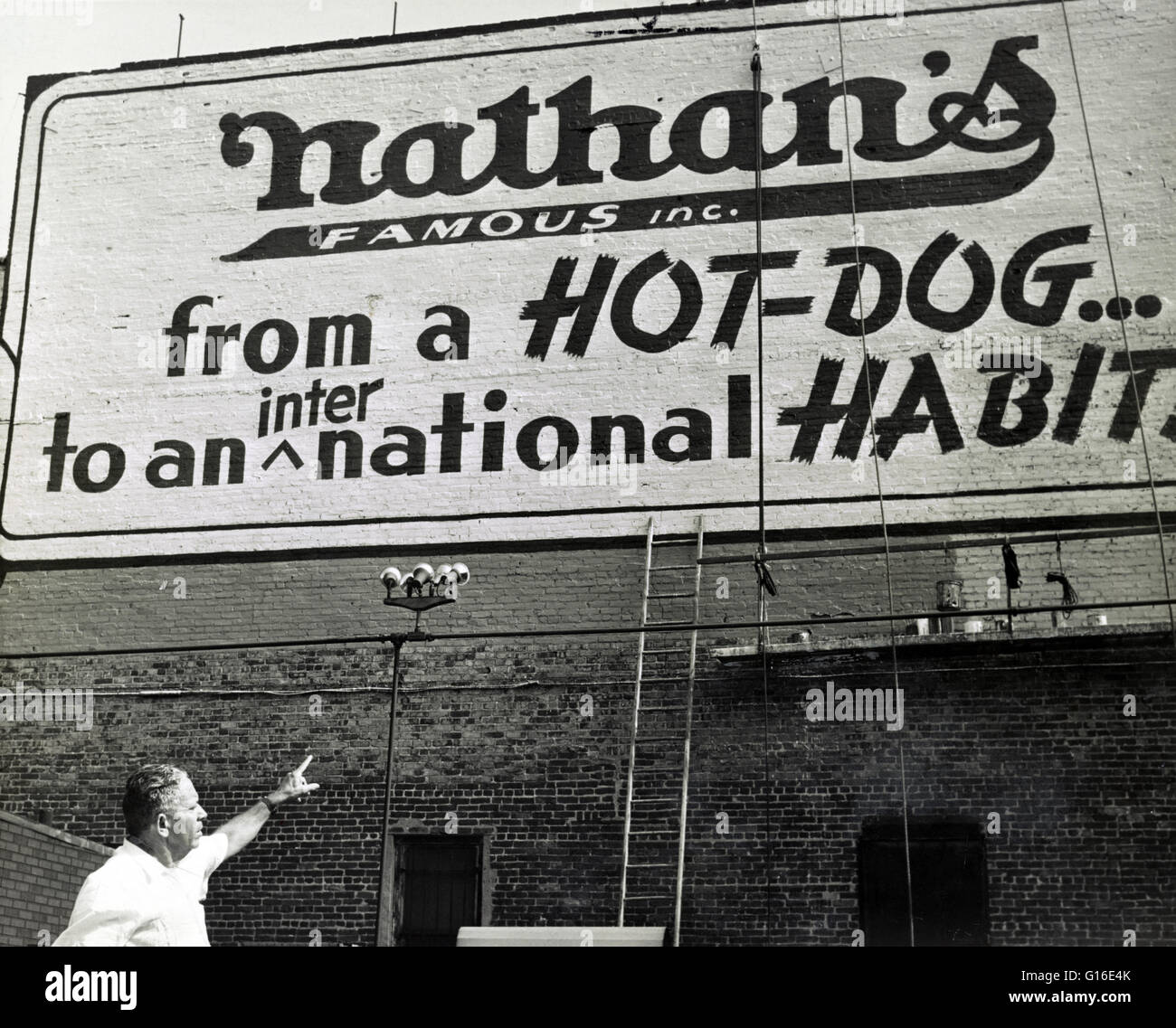 Entitled: 'From a hot dog to an international habit' showing Nathan Handwerker pointing to restaurant sign painted on outside wall.  Nathan's Famous, Inc. is an American company that operates a chain of fast food restaurants specializing in hot dogs. Nath Stock Photo