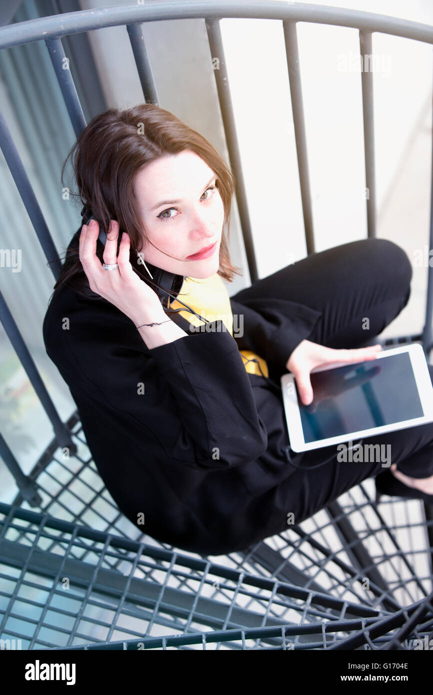 young woman sitting on stairs with headphones and holding an ebook Stock Photo