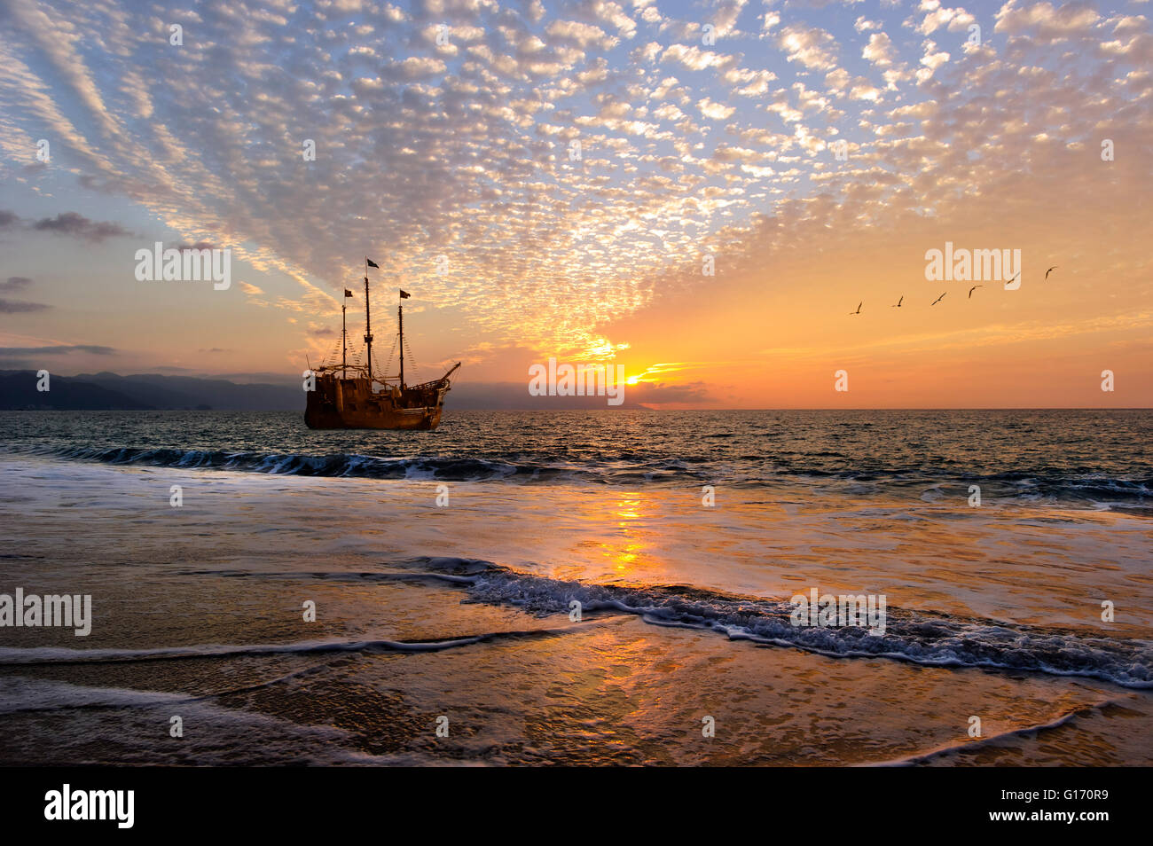 Ship sunset is an old wooden pirate ship with full flags as the sun sets on the ocean horizon in a colorful sunset sky. Stock Photo