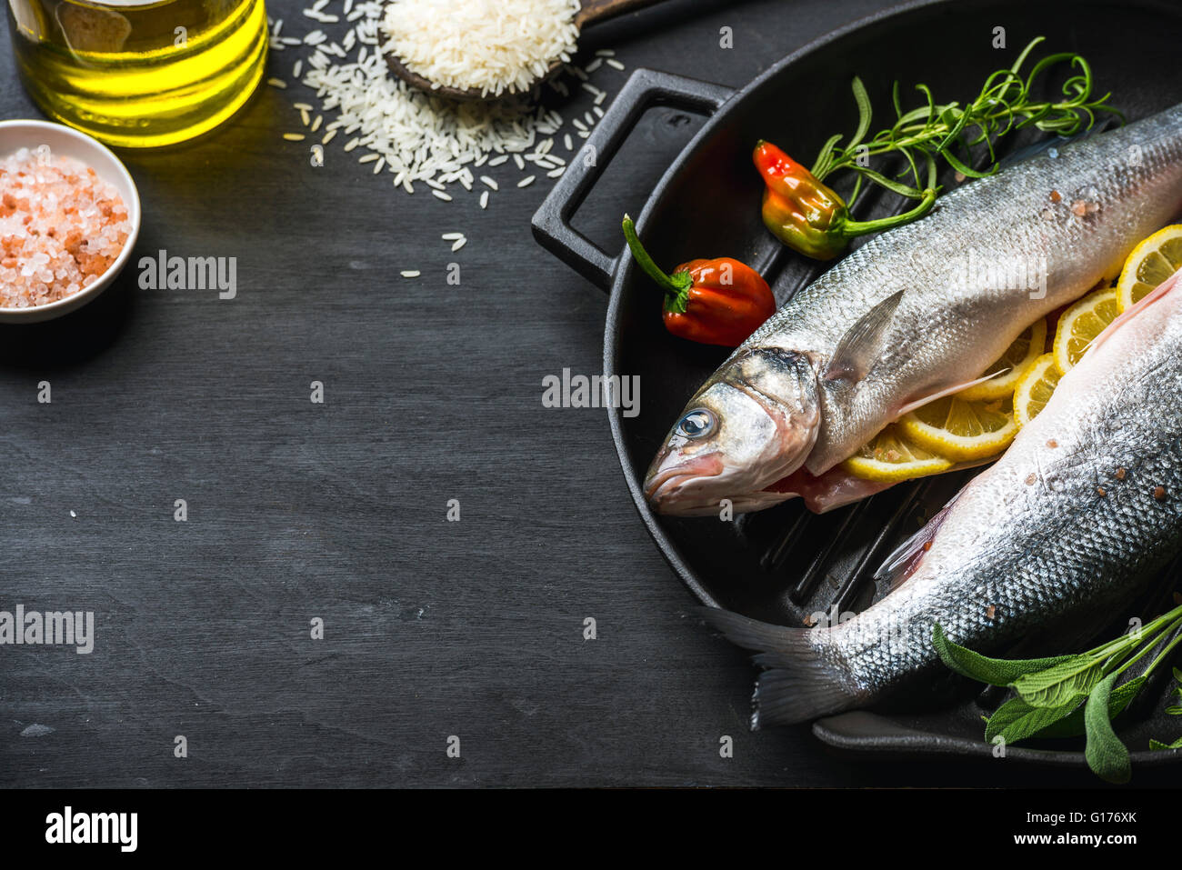 Raw uncooked seabass fish with herbs, spices and rice in cast iron cooking pan on black wooden background. Copy space, selective Stock Photo
