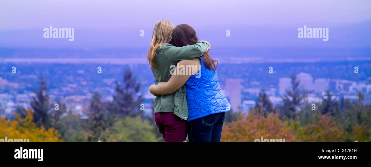 Two young women, standing on hillside, hugging, looking at view, rear view Stock Photo